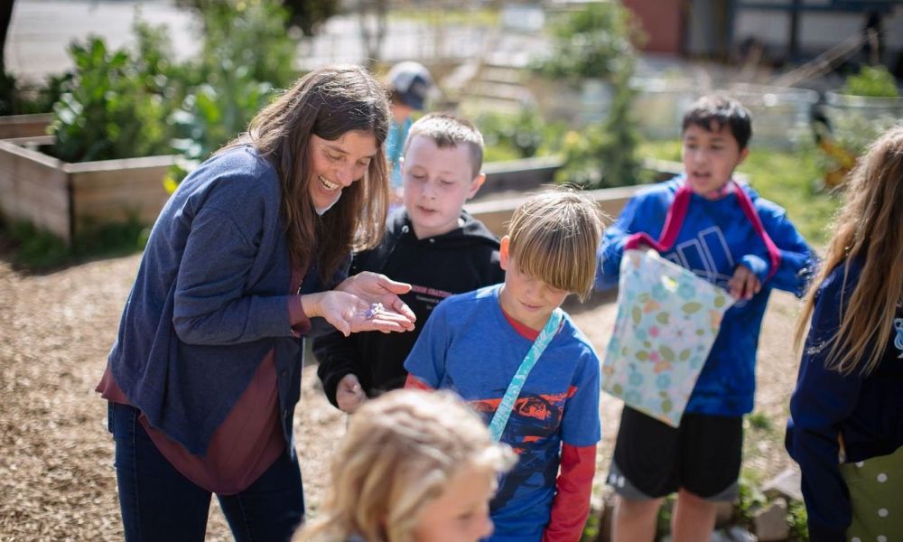 Students and a teacher learning in the garden. The teacher is holding something in their hand, showing it to the students.