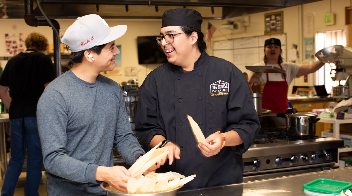 Two students standing at a metal kitchen work bench, smiling at each other. They have a bowl of corn husks that they are reaching into to.