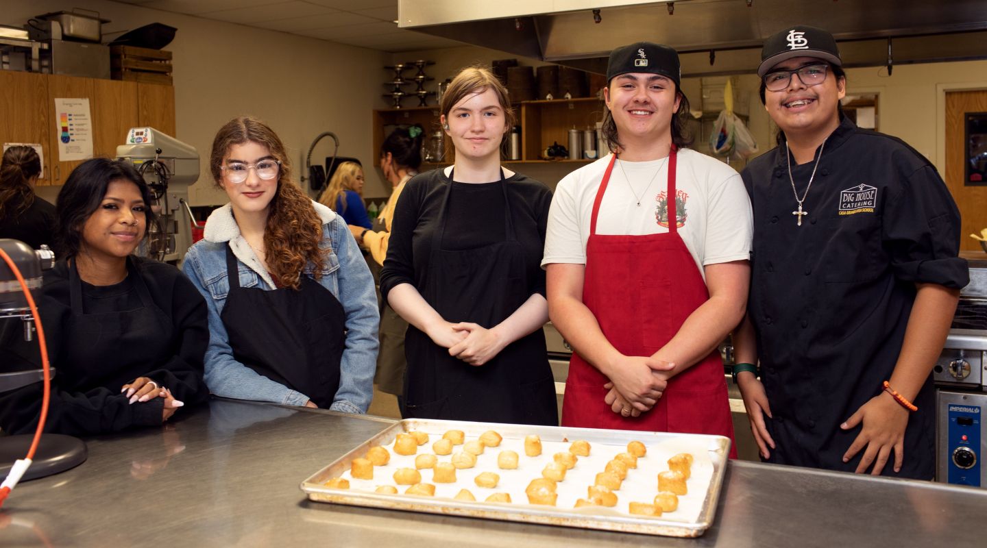 A group of Casa Grande students standing behind a metal kitchen table, smiling for the camera. In front of them is a baking tray filled with baked goods.