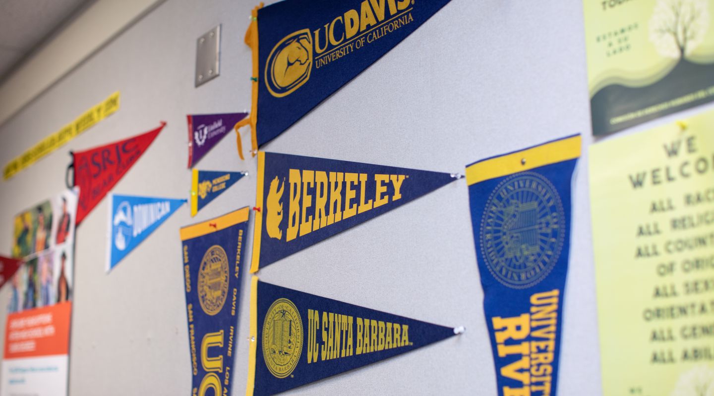 An image of college flags hanging on a wall in a classroom. The flags that are center in frame are "UC Davis", "Berkeley", and "UC Santa Barbara".