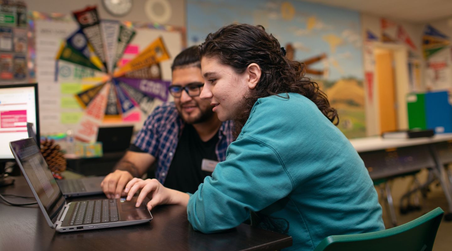 A student and teacher sitting at a desk and looking at a laptop together.