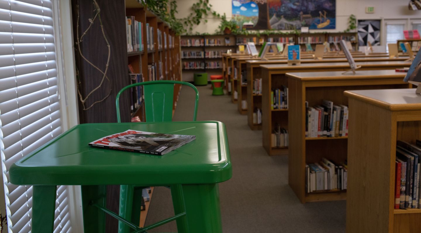 A view of a green metal table with a chair. Behind it is a view of many rows of wooden bookshelves filled with books.