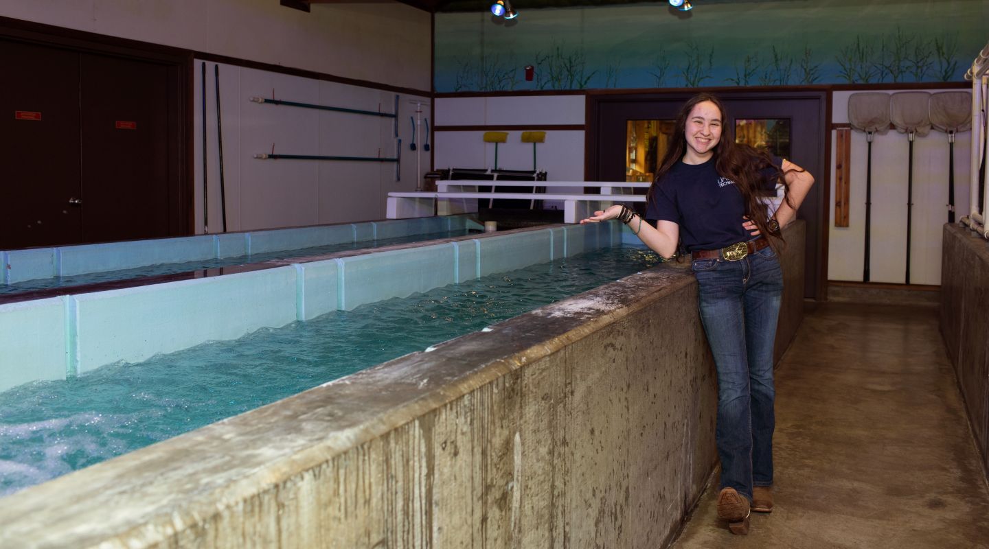 A Casa Grande High School student showing off the inside of the Casa Hatchery building. Next to the student, there is a large trough frilled with water.