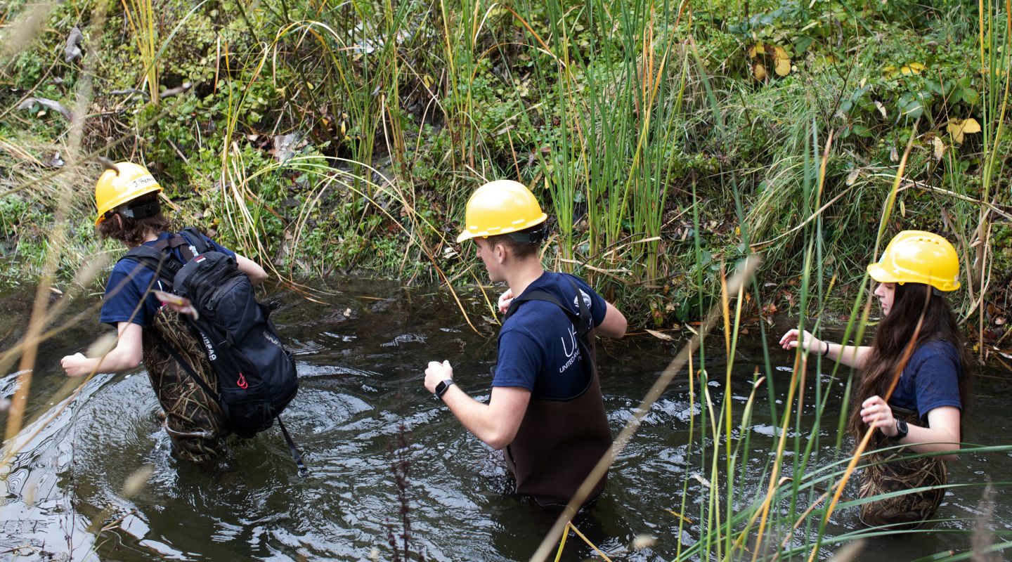 Three students wading through a waist-deep river wearing yellow safety hats.