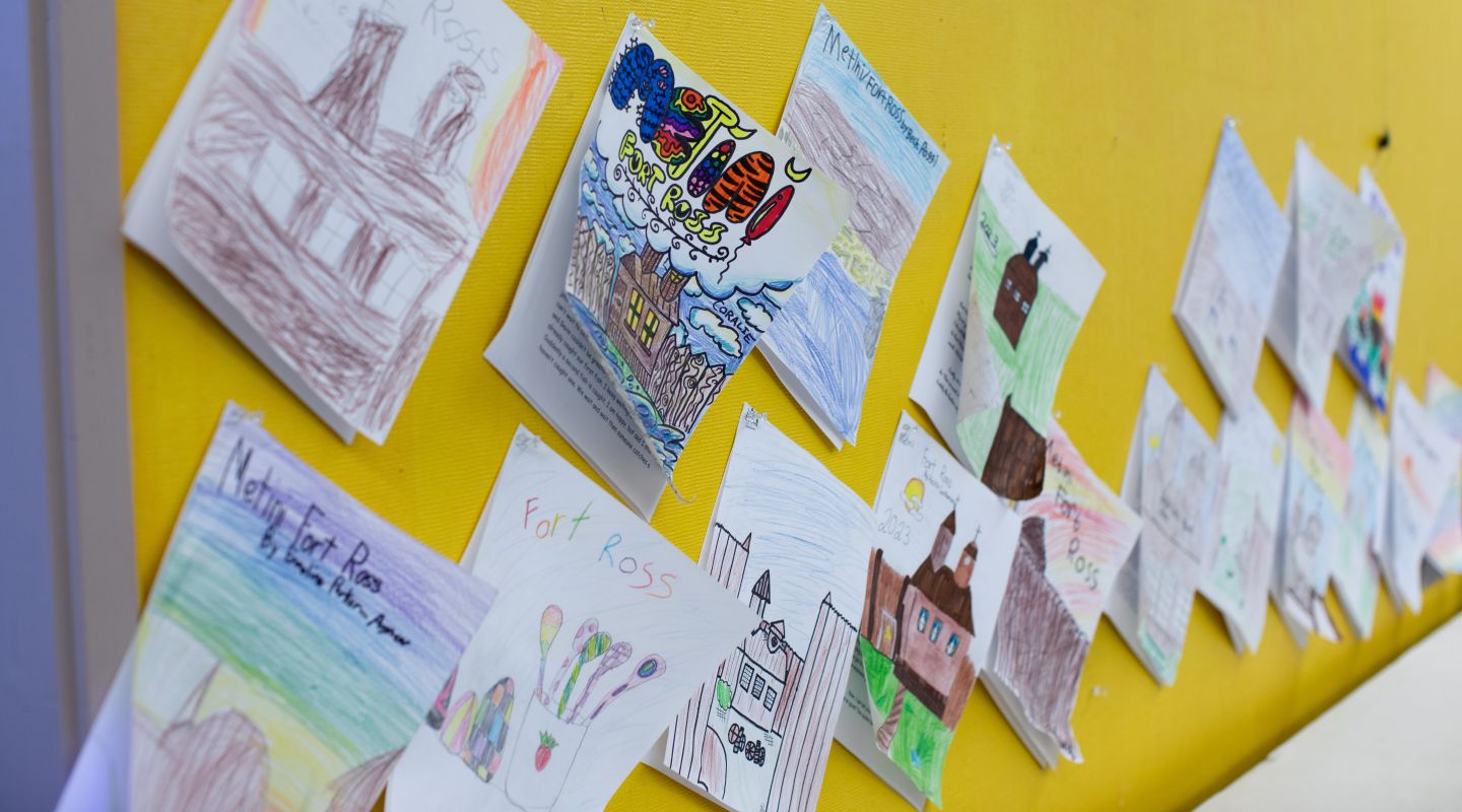 Multiple students' paper artwork of Fort Ross displayed on a yellow wall.