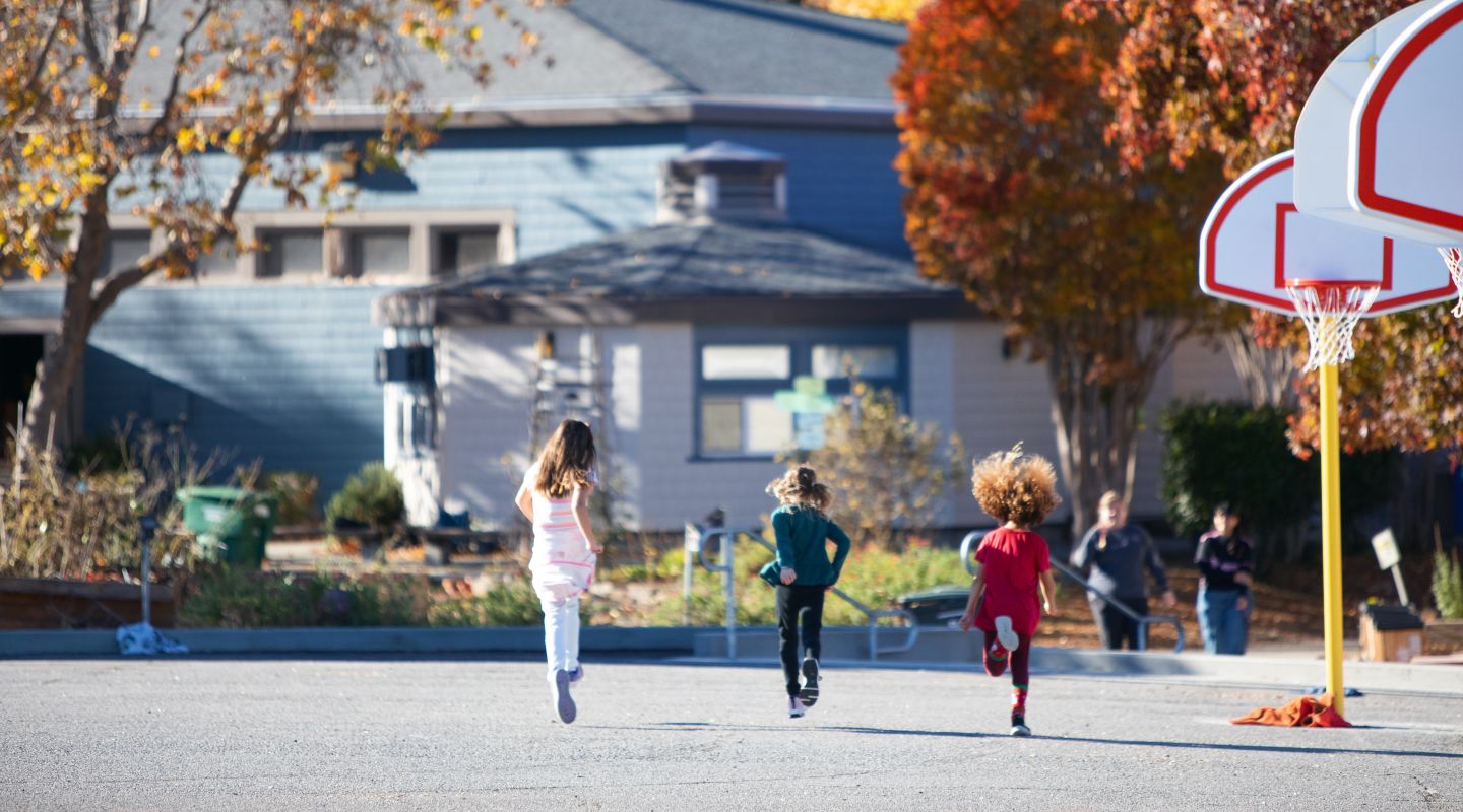 Three children in the distance, running away from the camera on an outdoor basketball court.