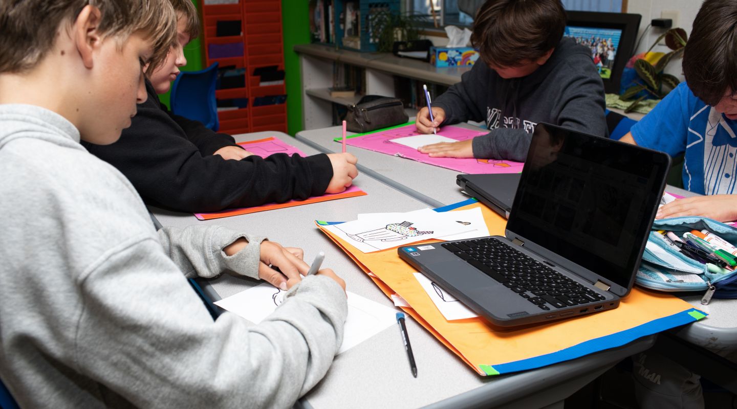 A student sitting at a desk, writing on a piece of paper, with a laptop open in front of them.