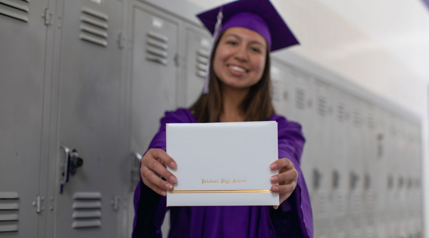 Petaluma High School student standing in front of gray lockers, wearing a purple graduation gown, and holding a diploma in front of the camera