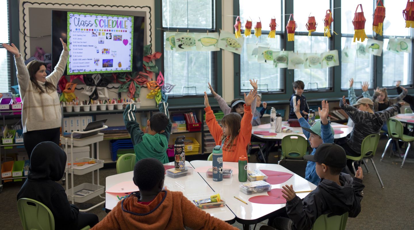 Penngrove Elementary students sitting at tables during class, and raising their hands in the air.