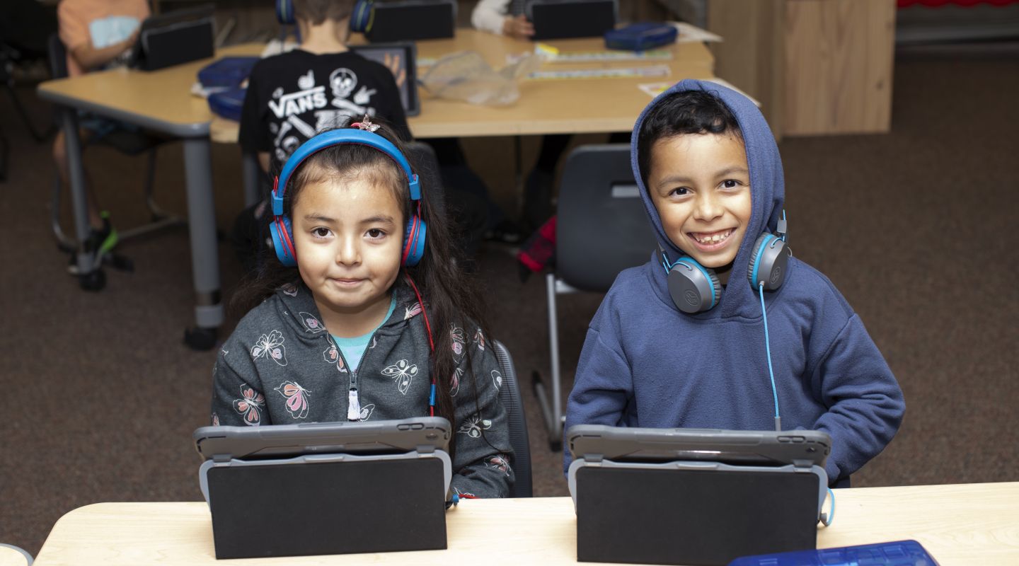 Two Penngrove students sitting at a desk, smiling for the camera. Both students are wearing headphones and using ipads.