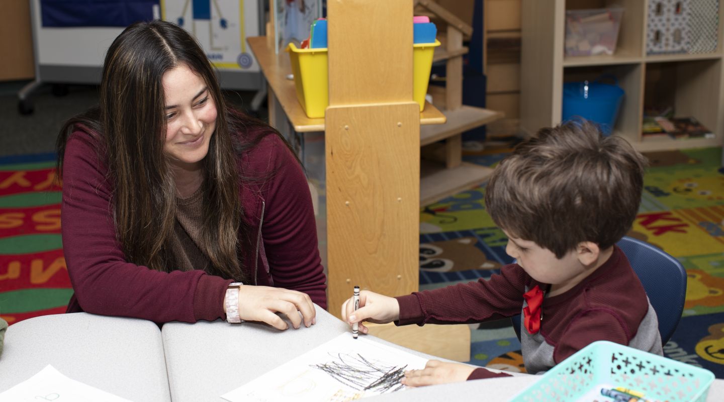 A student and a teacher sitting at a desk together. The child is drawing with a black crayon onto a piece of paper.