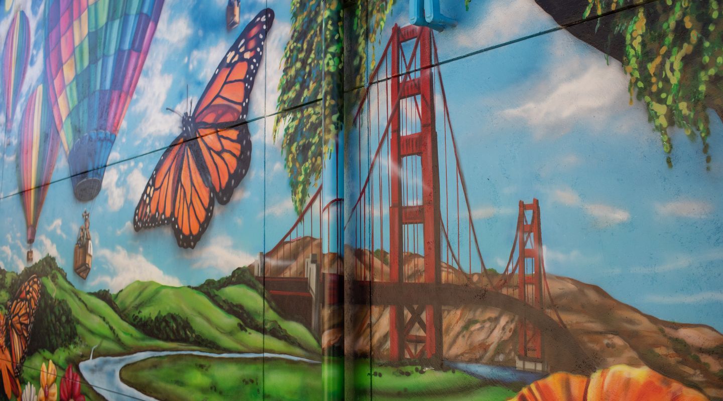 A mural on the outside of Penngrove Elementary that depicts a landscape scene including the golden gate bridge, an orange butterfly, hot air balloons, and a rolling hillside with a stream.