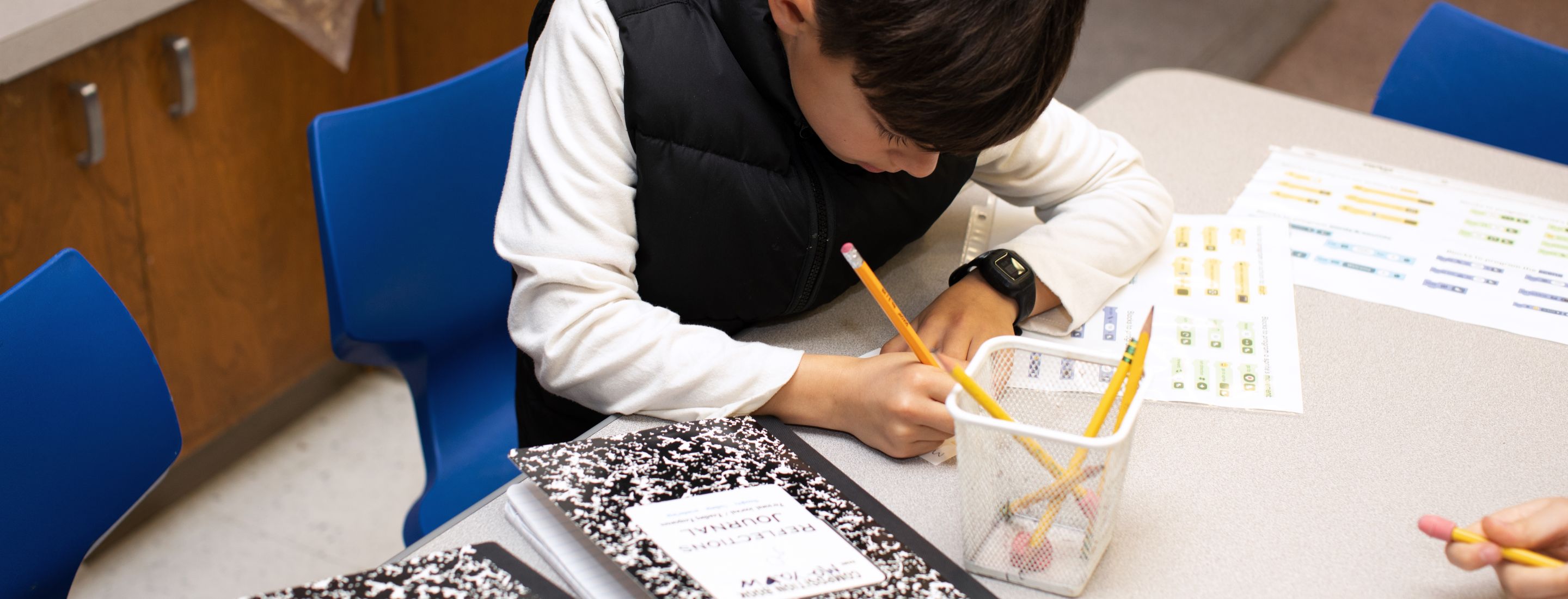 A student sitting and working at a desk with a pencil. In front of the student, on the desk, is a black and white composition notebook.